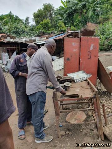 Rolle 1: Die Schraube, die wir für die defekte Stuhl-Rolle mitgebracht haben, wird erst mal an einem lose auf einem schief stehenden Tisch mit der bloßen Hand der Kopf weitgehend abgeschliffen. Damit er die Schraube zwischendurch abkühlen kann, hält er sie ab und zu in die Büchse mit Wasser...